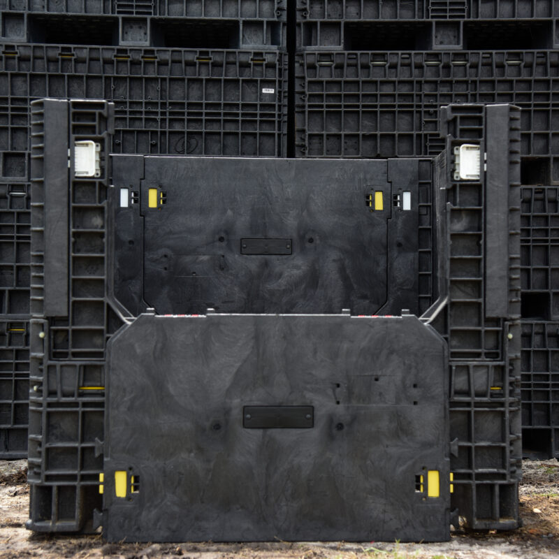Used black collapsible bulk container with its front access door lowered, pictured before stacked containers.